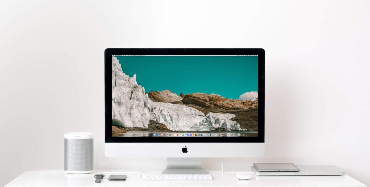 A collection of Apple products centered around an iMac on a white desk with a white wall.
