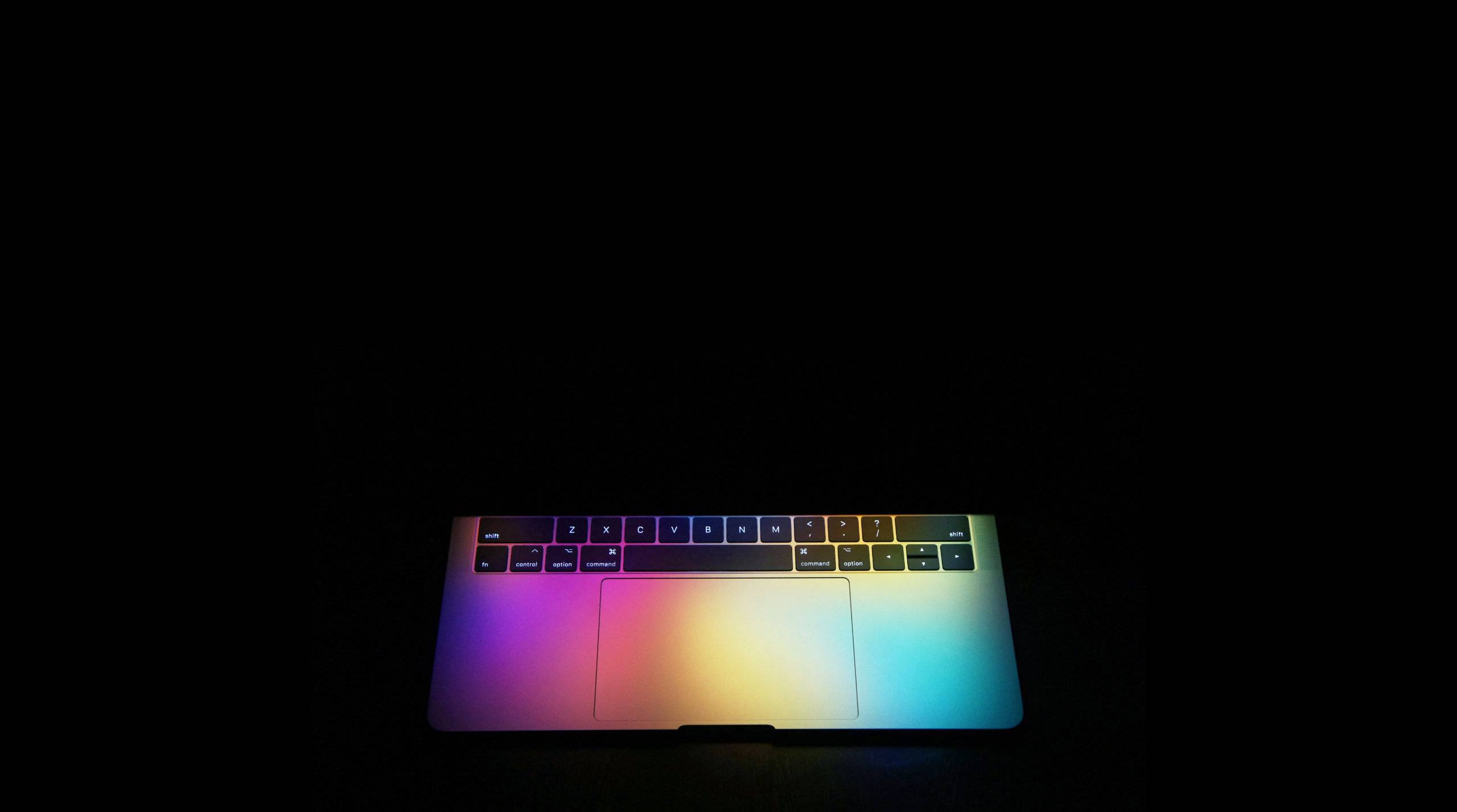 A MacBook Pro partially opened on a black background, a colourful glow cascades out across the aluminium.