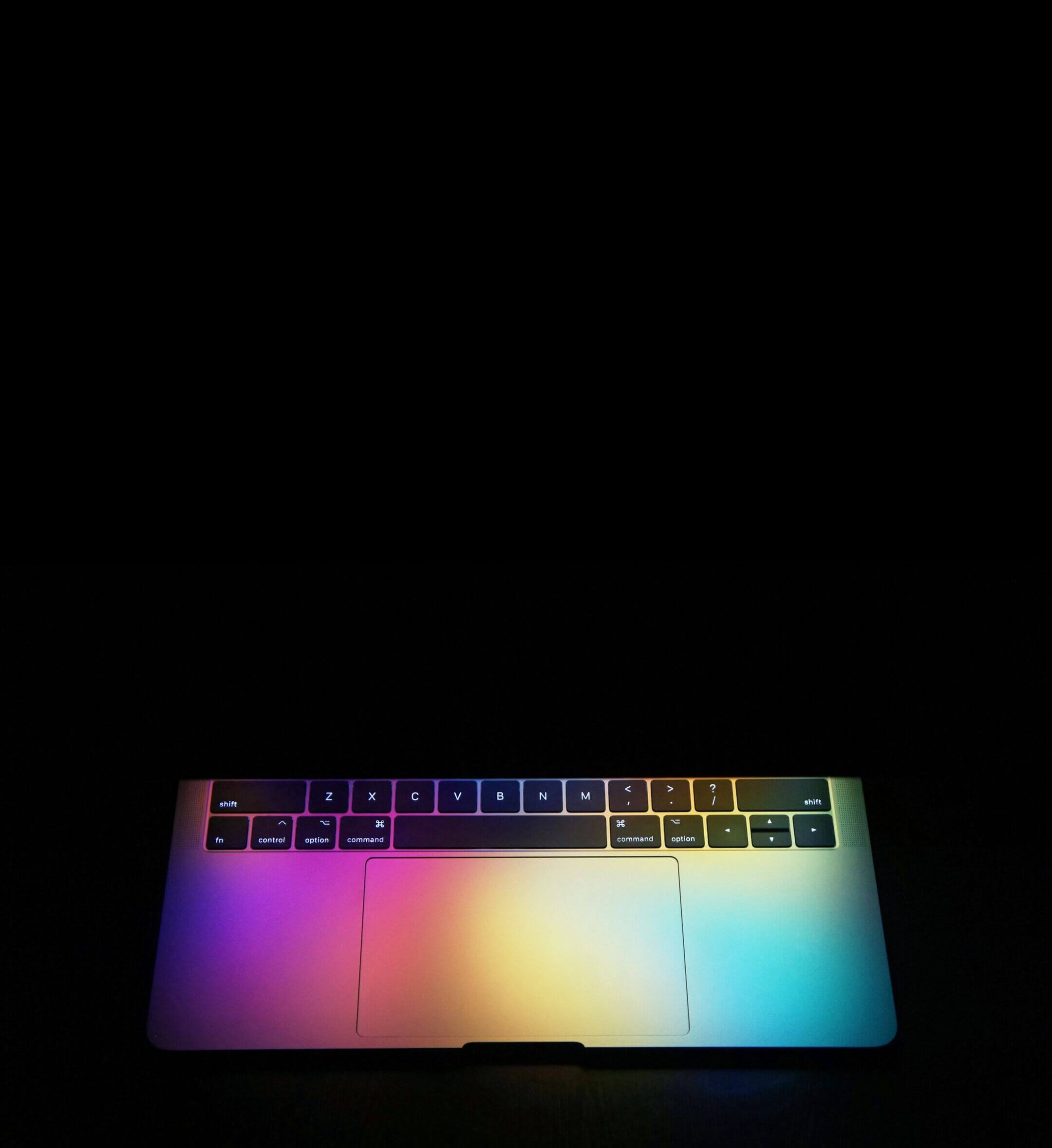 A MacBook Pro partially opened on a black background, a colourful glow cascades out across the aluminium.