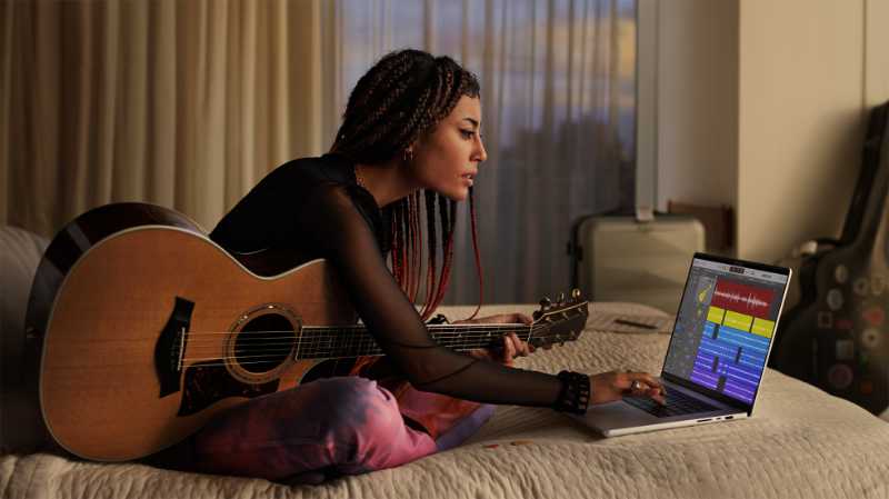 A woman sits on her bed, guitar on her lap, reaching towards a MacBook Pro running Logic Pro.
